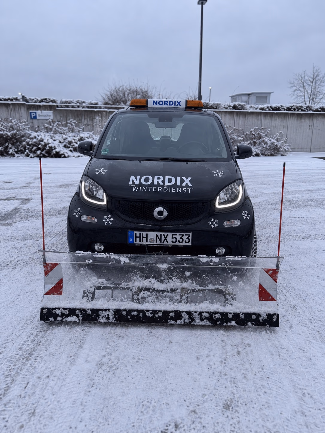 Schwarzer Smart Fortwo von Nordix Winterdienst mit Kennzeichen HH·NX 533, Räumschild und Rundumleuchte auf einem geräumten, verschneiten Parkplatz.